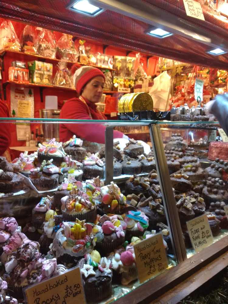 A woman sells colorful, decorated muffins and rocky road at a market stall in the UK or Ireland.