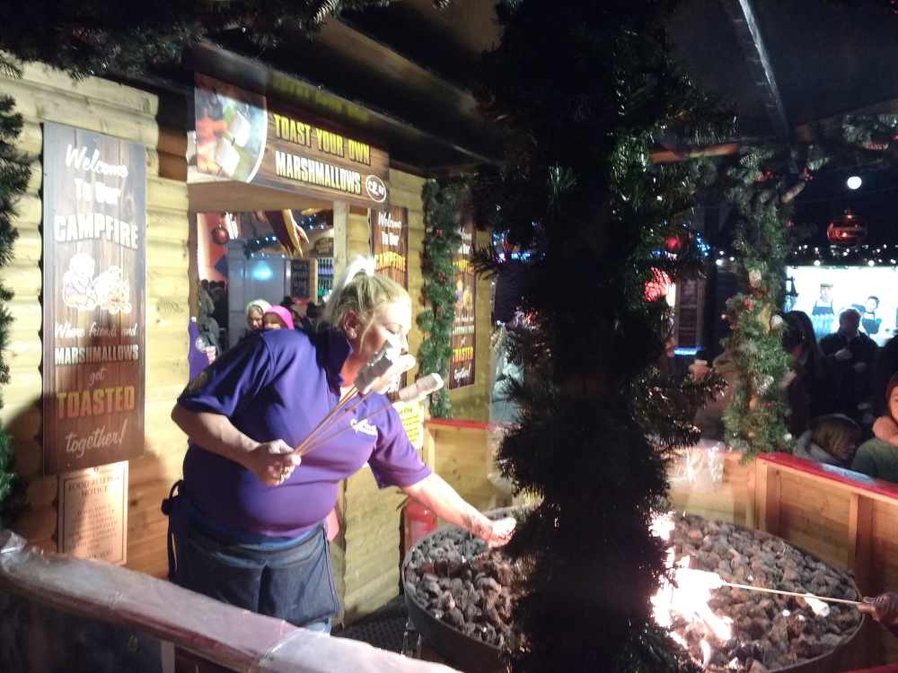 A woman roasts marshmallows over a fire pit at a bustling European Christmas market.