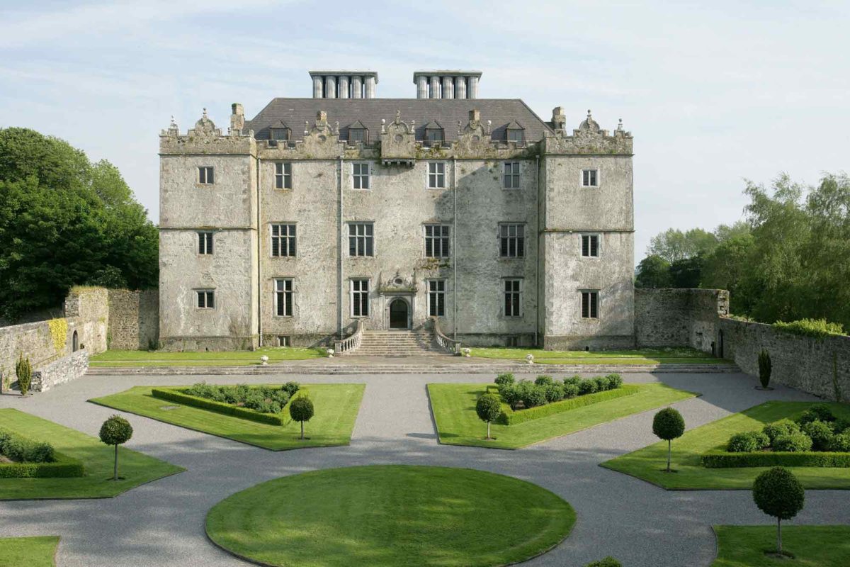 A large stone mansion with a symmetrical facade stands behind a formal garden with gravel paths and manicured hedges.