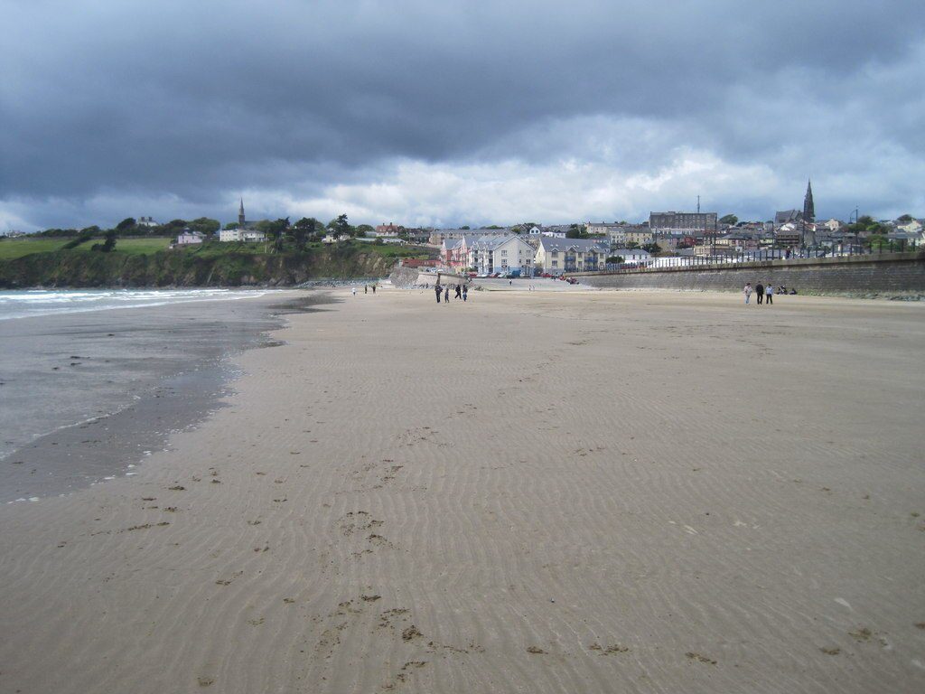 People walk along a wide, sandy beach beneath a cloudy sky, with a town and stone wall visible in the distance.