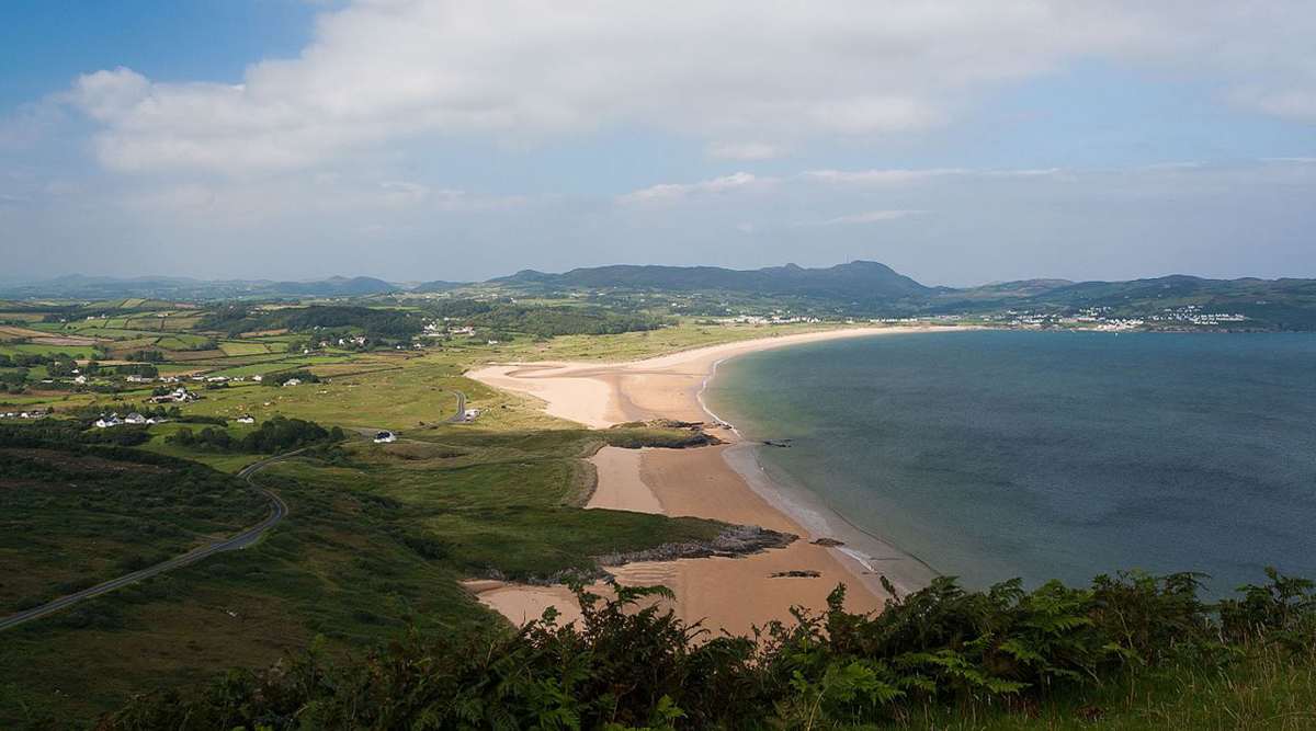 A wide sandy beach curves along a coastline bordered by green fields, hills, and a road under a partly cloudy sky.