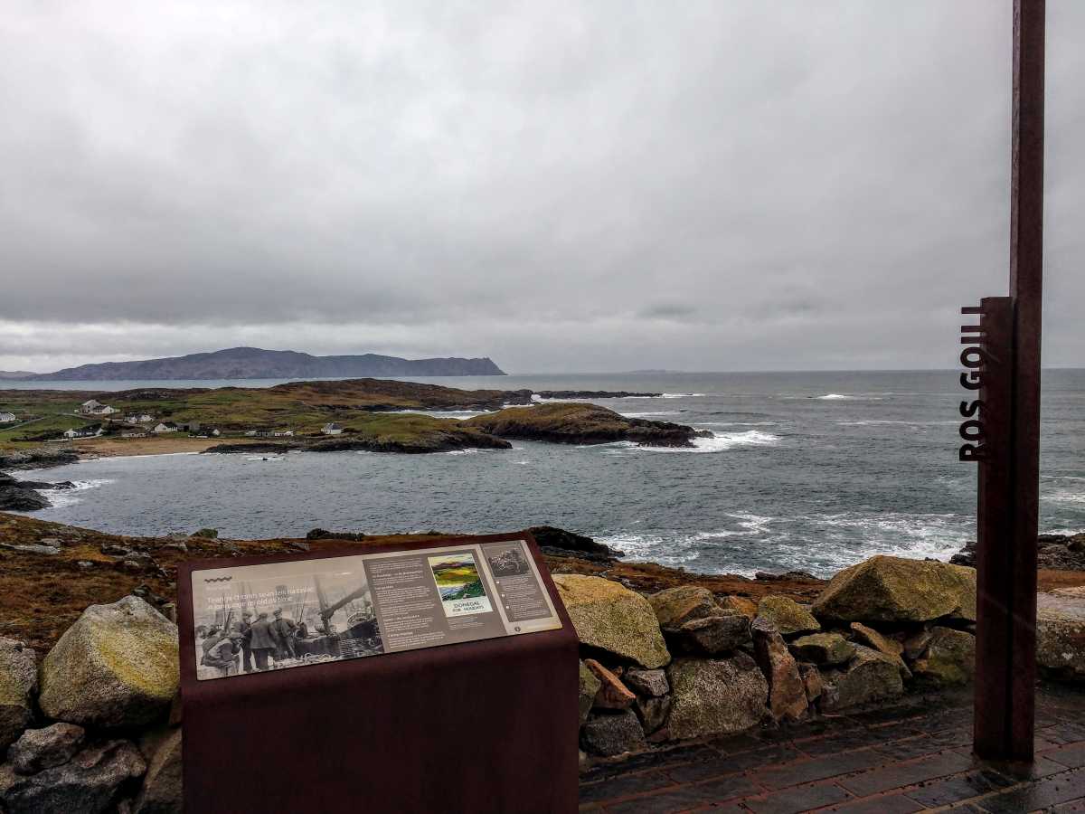 An information sign stands on a rocky coast overlooking a bay with a distant headland under a cloudy sky.