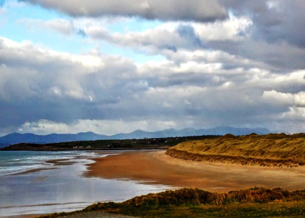 A sandy beach meets the ocean under a cloudy sky with distant mountains on the horizon.
