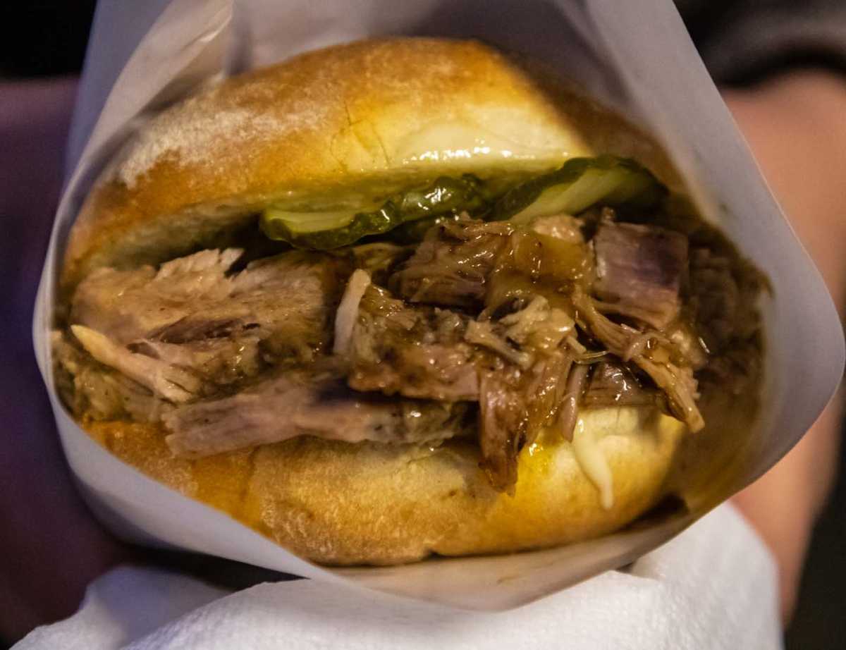 A pulled pork sandwich with pickles and gravy is held in a white paper wrapper.