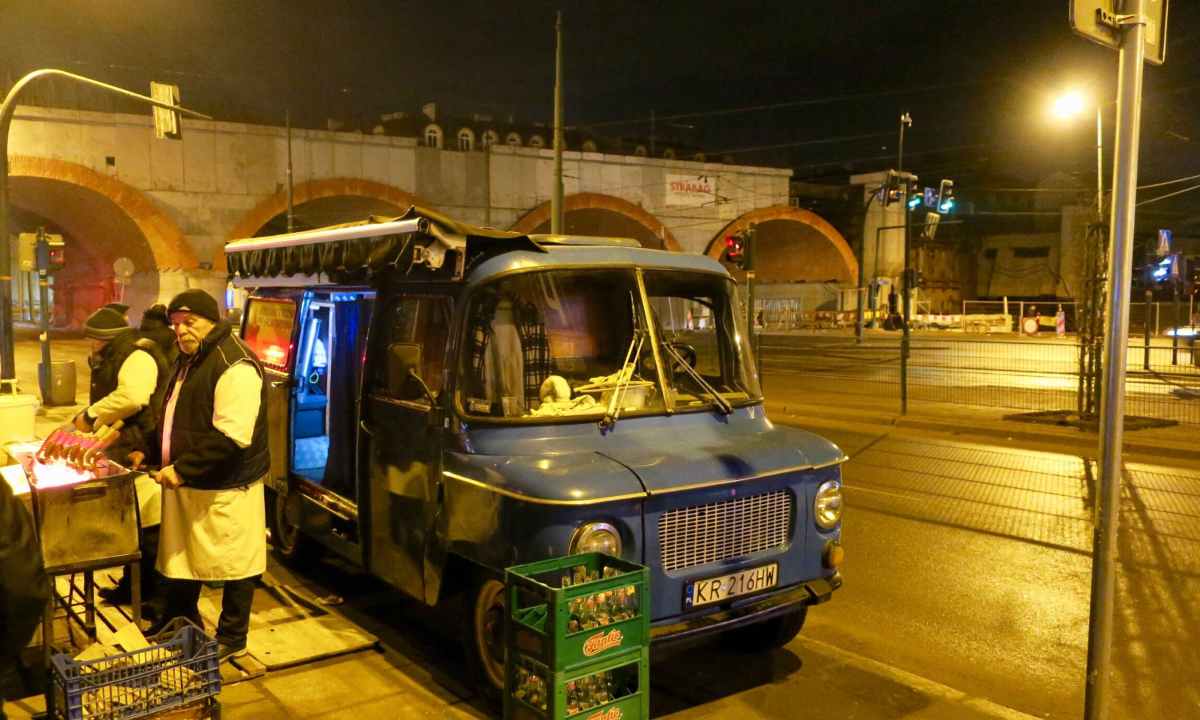 A man in a white apron grills sausages next to a blue food truck parked on a city street at night.