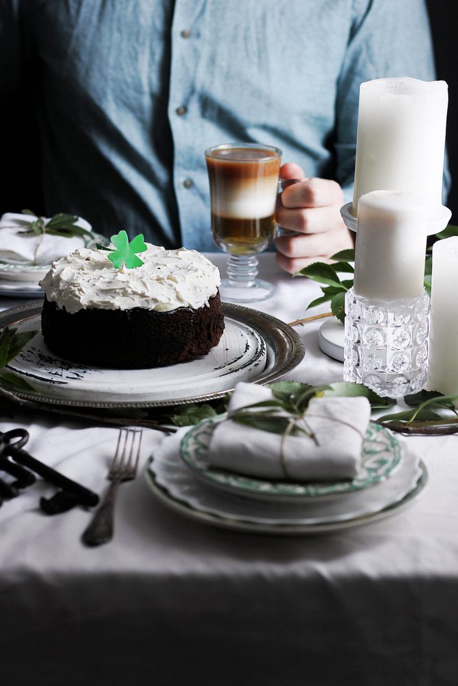 A person holds an Irish coffee next to a shamrock-topped cake and candles on a festive table.