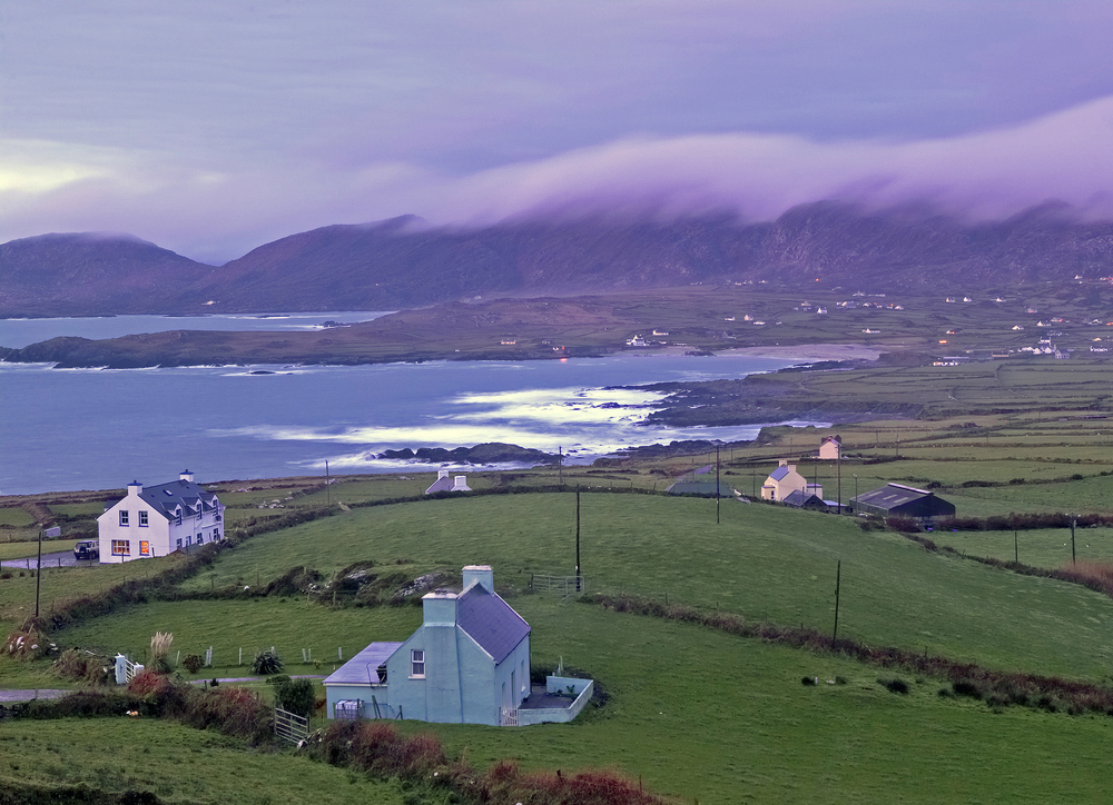Ring of Kerry coastline in Ireland with green fields, scattered houses, and misty mountains under a purple sky.