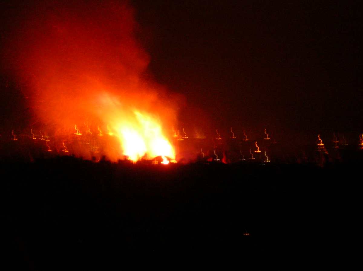 Large orange and yellow fire with smoke burns brightly at night over blurry distant lights.