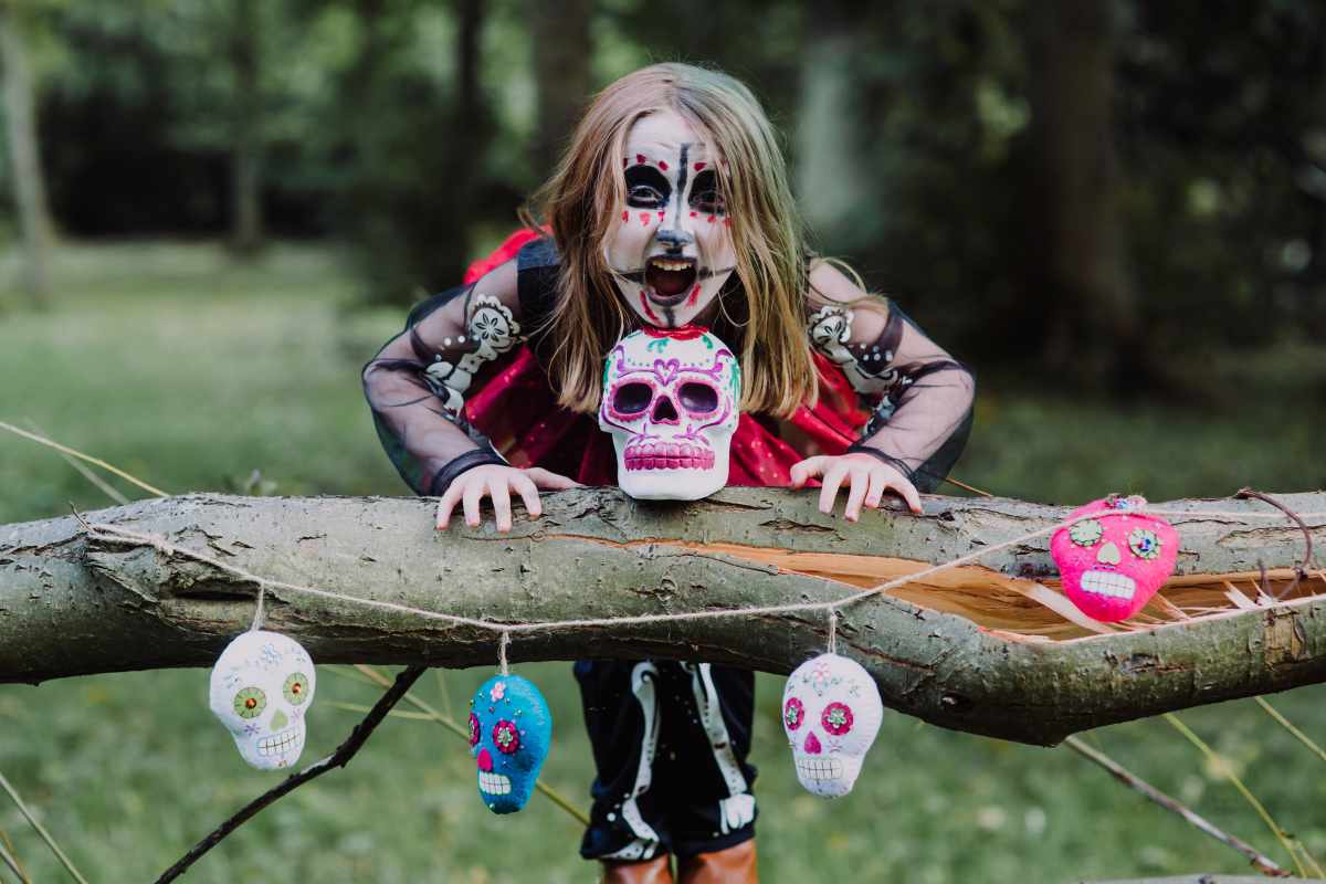 A child with sugar skull face paint and costume leans over a log with colorful skull decorations.