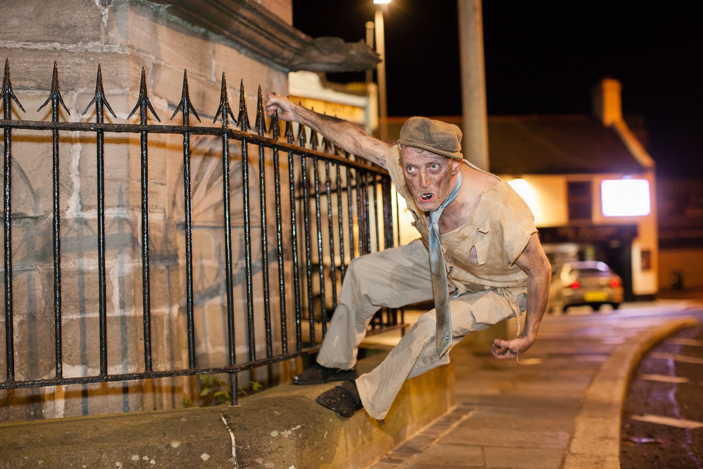 A ghoulish man in tattered clothes and a flat cap climbs a spiked fence on a dark street.