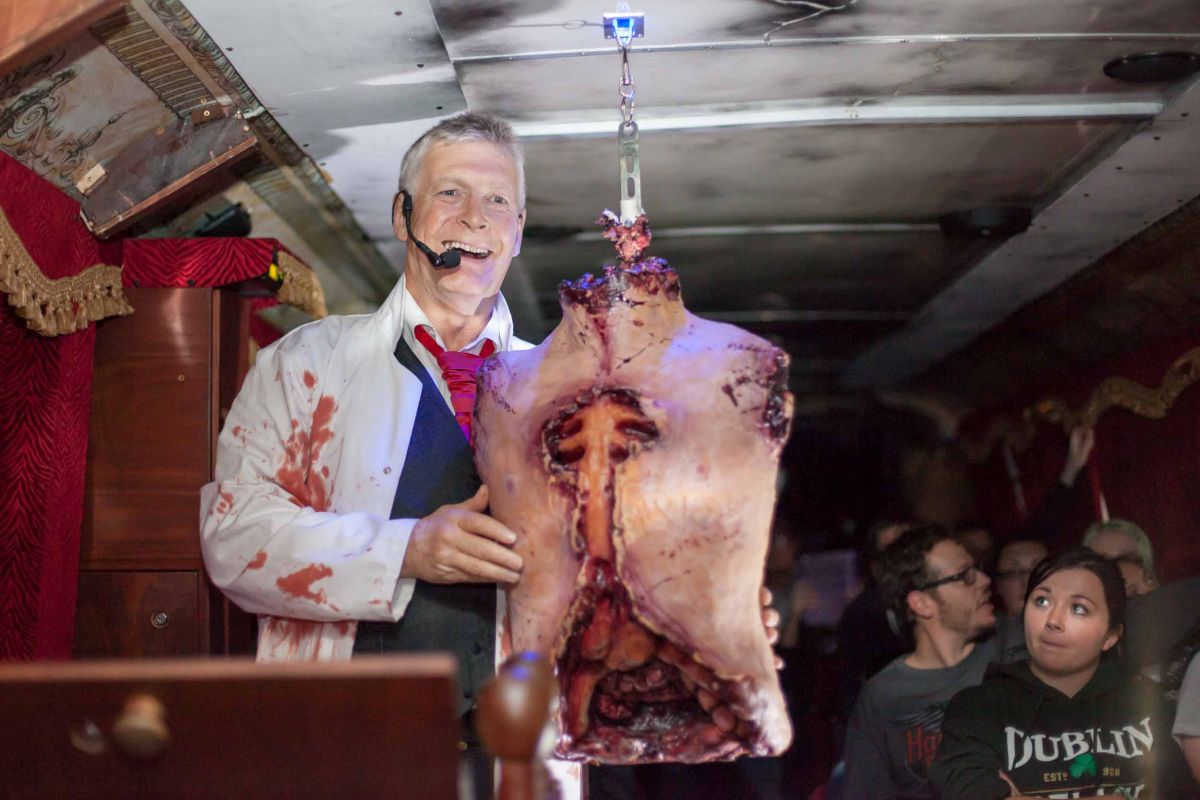 Smiling actor in a bloody lab coat presents a gruesome prop to an audience in Dublin.