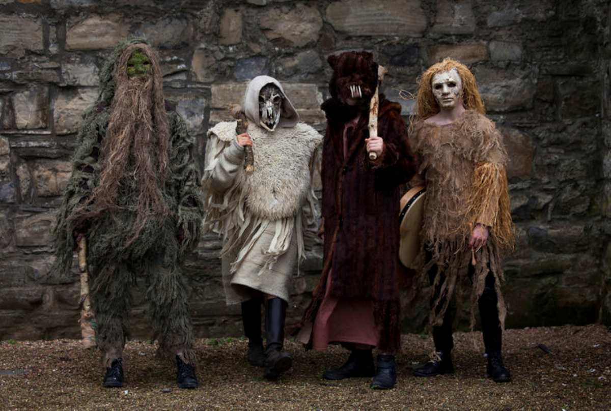 Four people in elaborate folk costumes, including a ghillie suit and a bodhrán, stand against a stone wall.