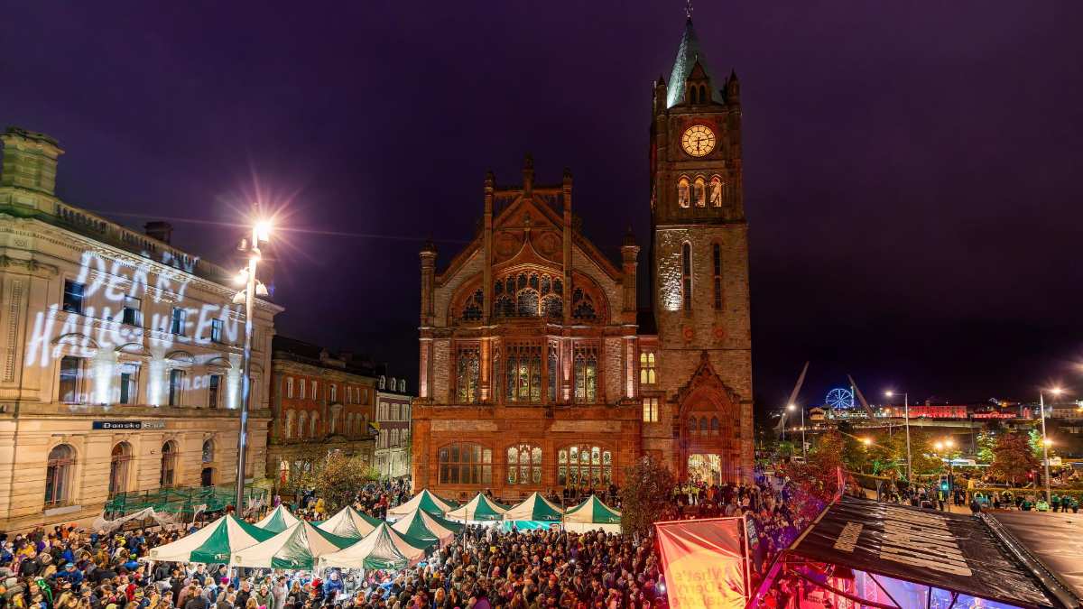 Derry Guildhall illuminated at night during a bustling Halloween festival with crowds and market stalls.