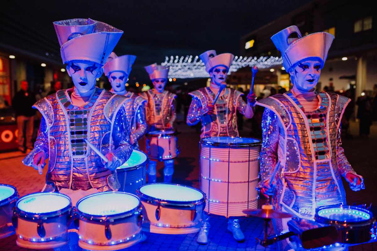 Group of performers in futuristic sequined costumes with white face paint play light-up drums at a night event.