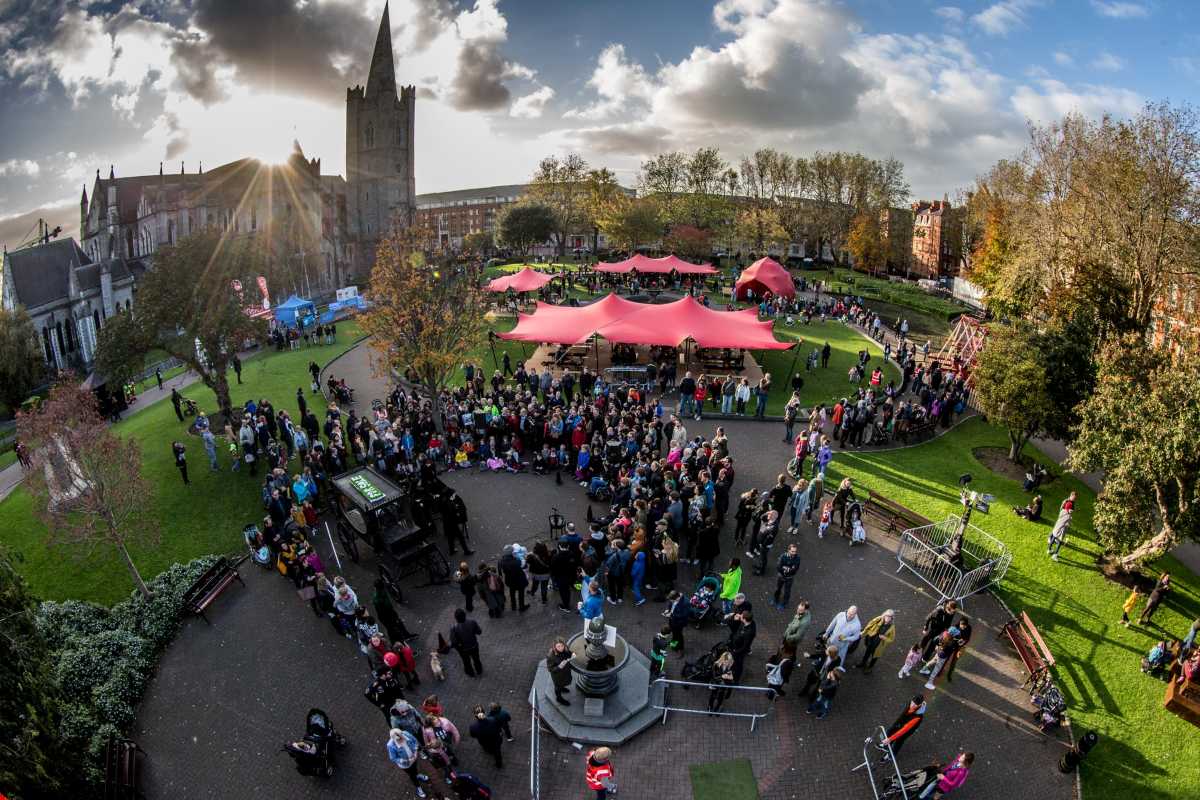 Sunlight beams behind St. Patricks Cathedral, Dublin, as a large crowd enjoys a park event with red tents.