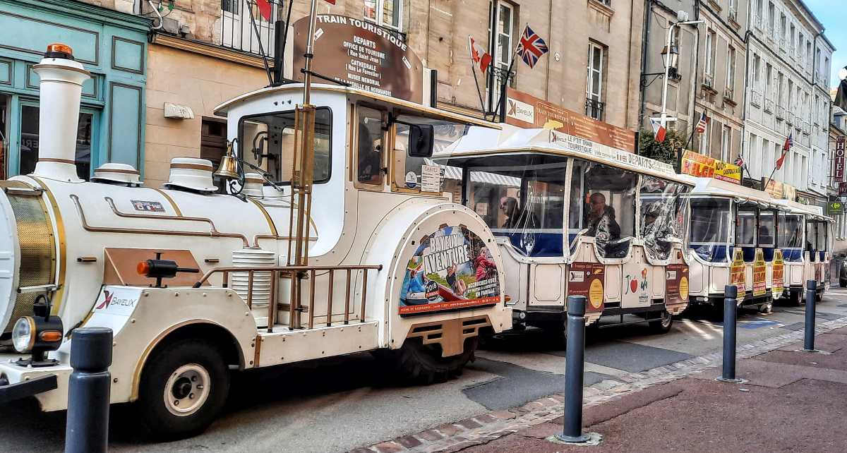 White tourist train parked on a street in Bayeux, France, with historic European buildings.