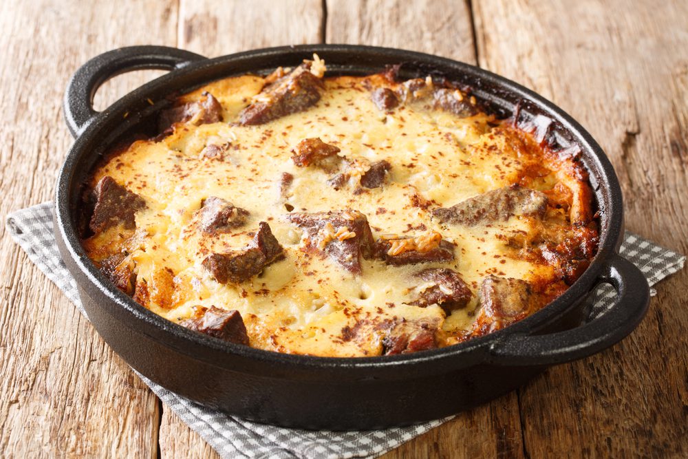A baked dish with meat chunks and a golden-brown  crust sits in a black cast-iron pan on a wooden surface.