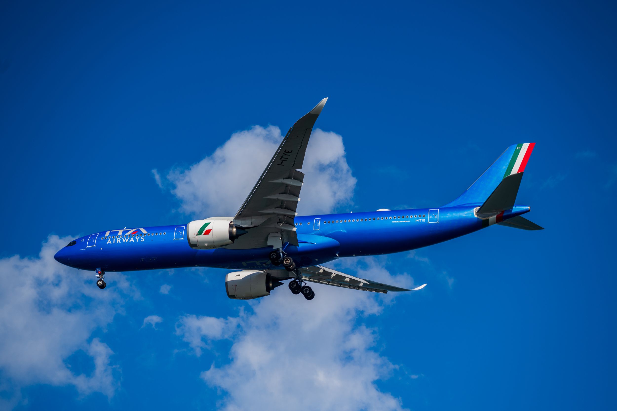 A vibrant blue ITA Airways airplane captured mid-flight against a clear blue sky with clouds.