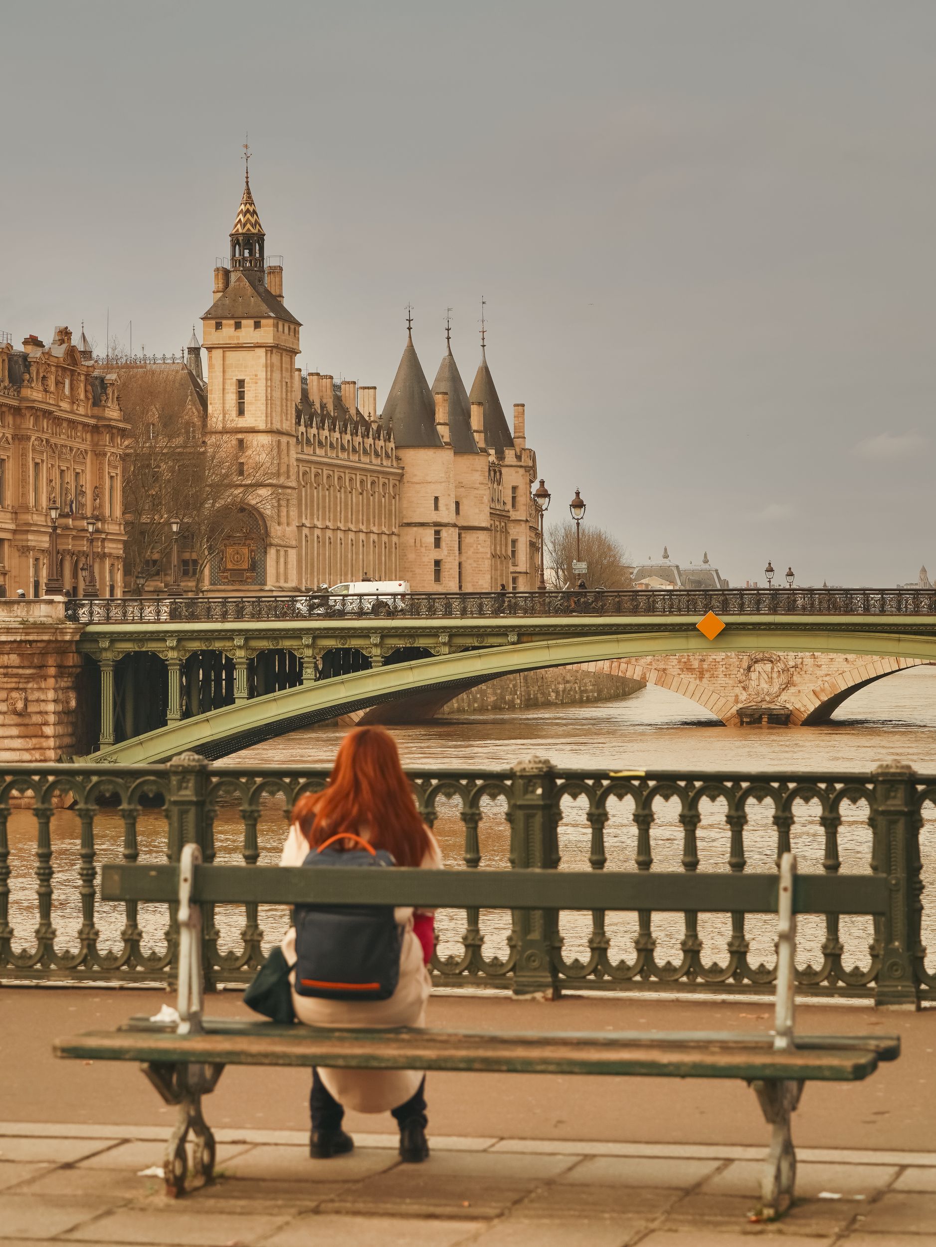 A woman sits on a bench overlooking the Seine River with the Conciergerie in the background.