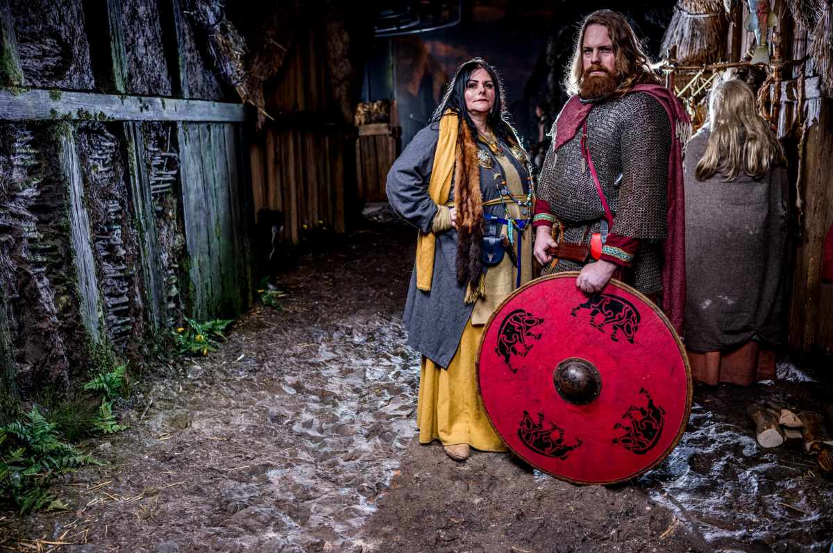 Two people in historical-style clothing stand on a dirt path next to a red shield with black animal designs.