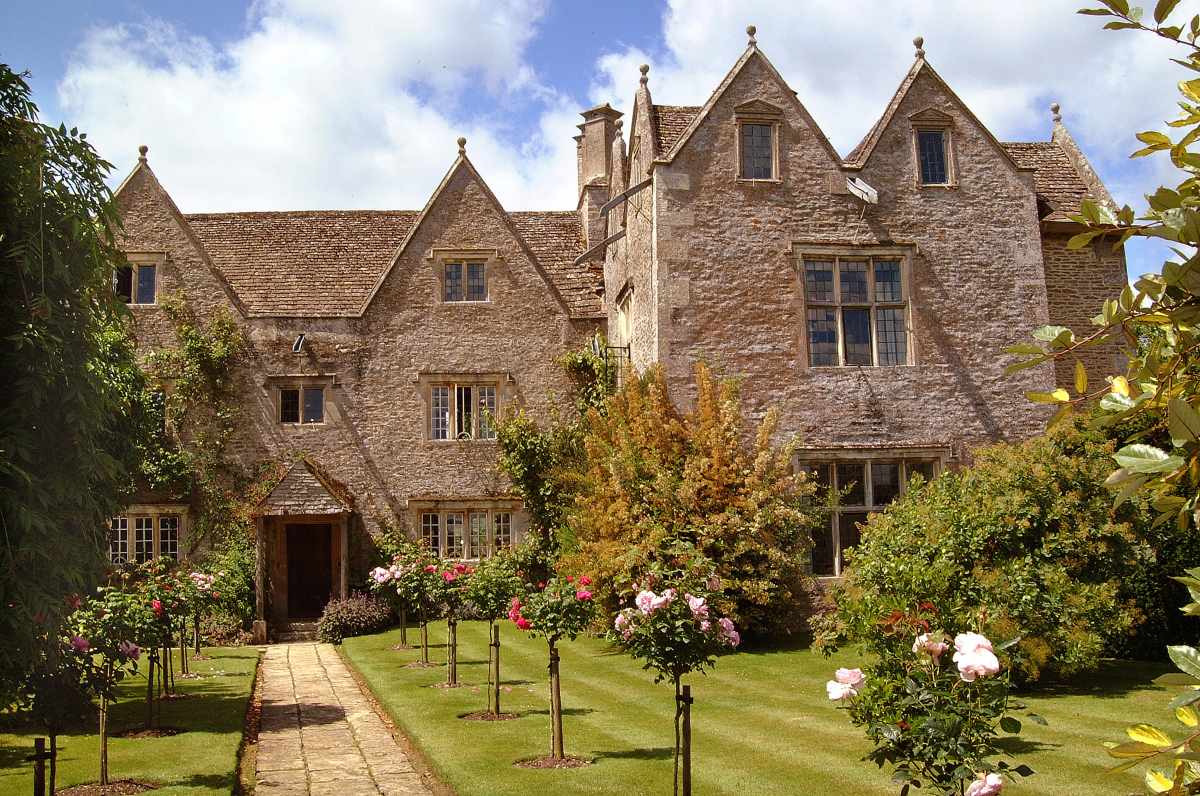 A stone manor house with multiple gables sits behind a garden path lined with rose bushes and a manicured lawn.