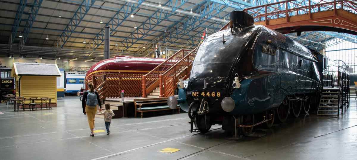 An adult and a child walk past a large, dark blue steam locomotive inside a spacious museum hall.