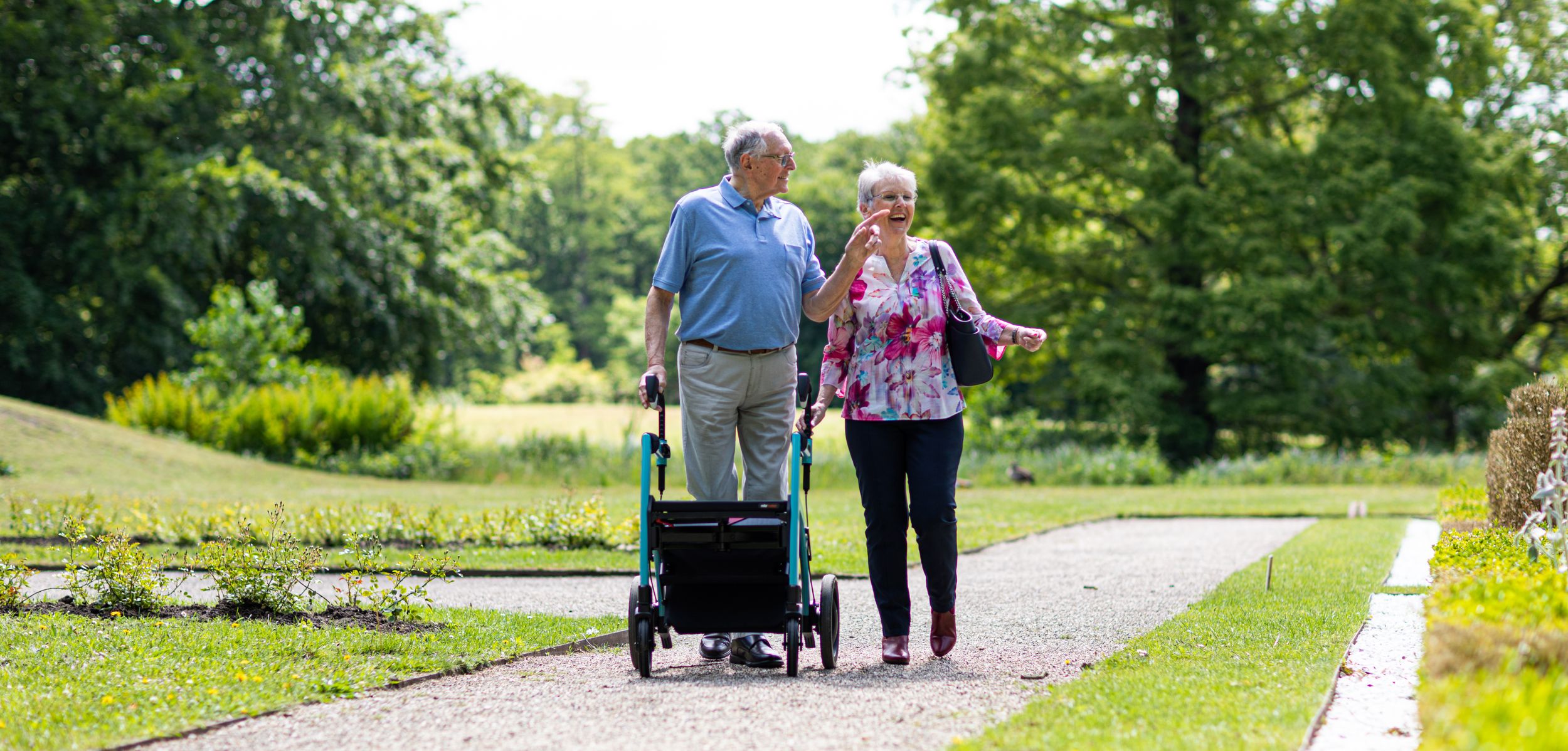 Happy elderly couple walks on a park path, the man using a rollator, surrounded by green trees.