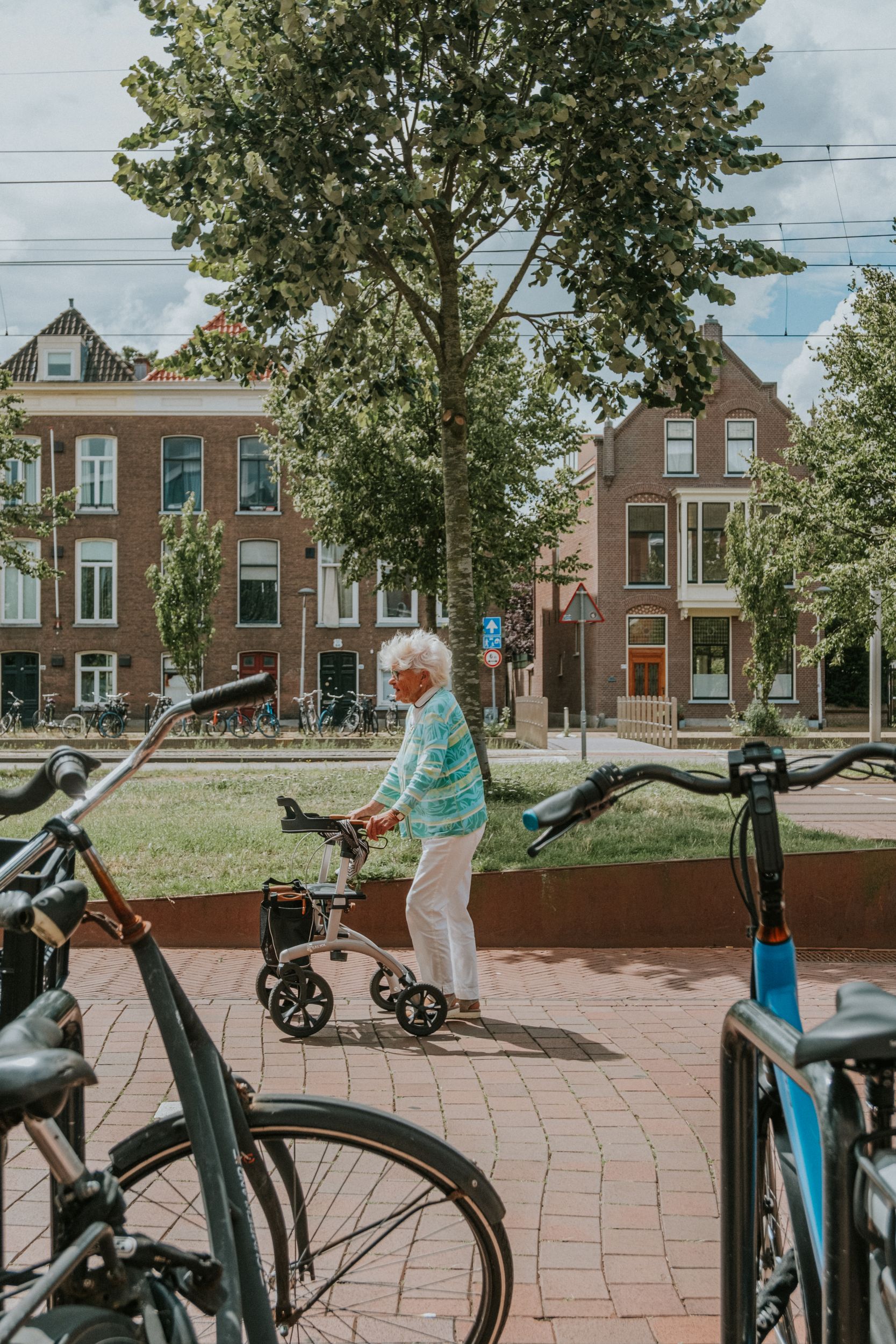 Elderly woman with a walker on a brick street in a Dutch city, with gabled brick houses and parked bicycles.