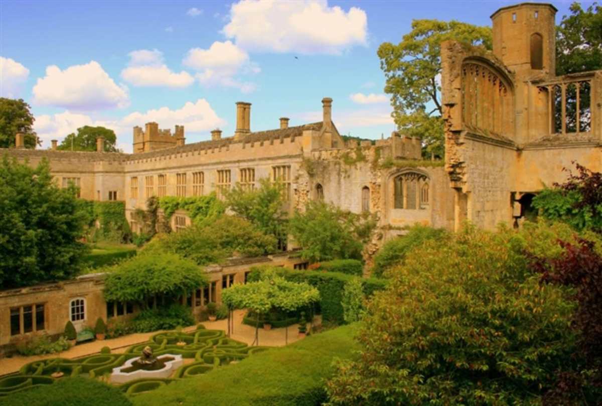 A stone building with a partially ruined tower overlooks a formal garden with manicured hedges and a central fountain.