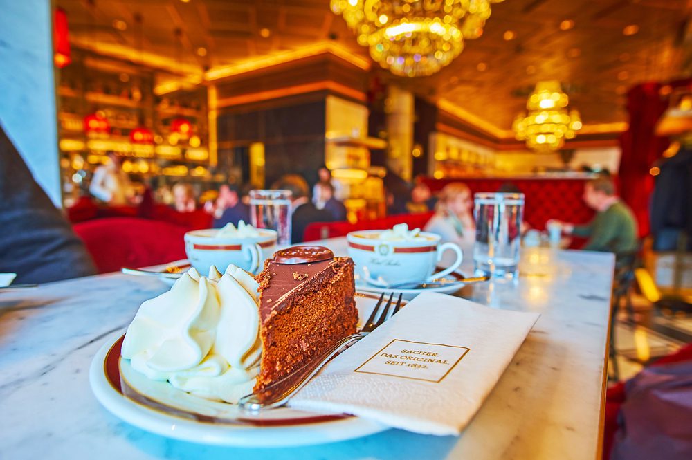 A slice of chocolate cake with whipped cream sits on a plate next to a napkin and coffee cups in a dimly lit restaurant.