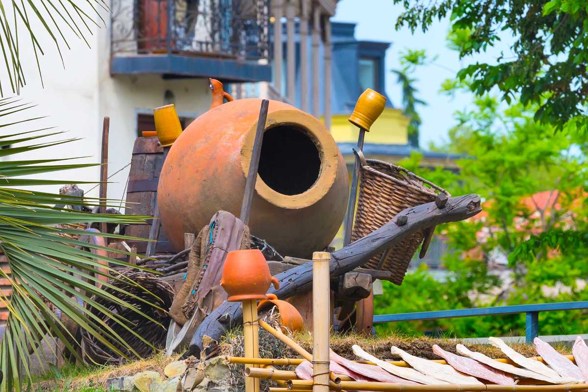 A large clay vessel sits among wooden barrels, baskets, and smaller pots in an outdoor setting with palm fronds.
