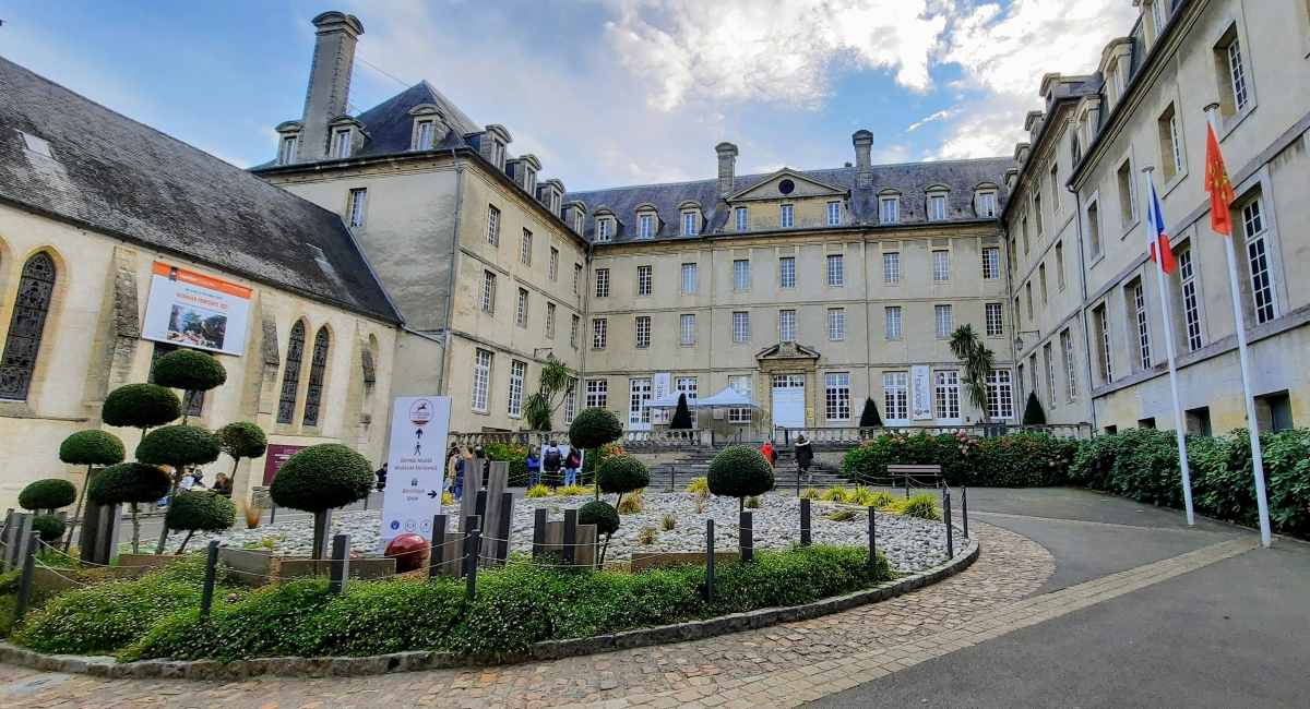A large stone building with a courtyard features manicured topiary trees, a gravel garden, and a paved path.