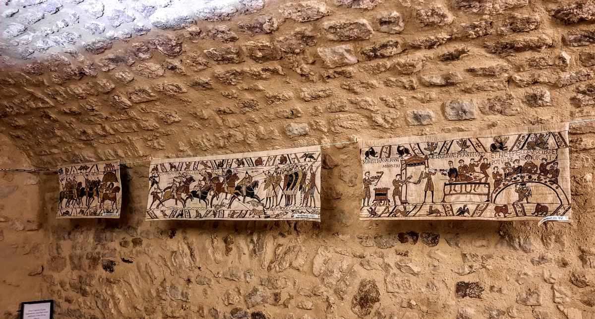 Three embroidered panels depicting historical scenes are hung on a rough stone wall.