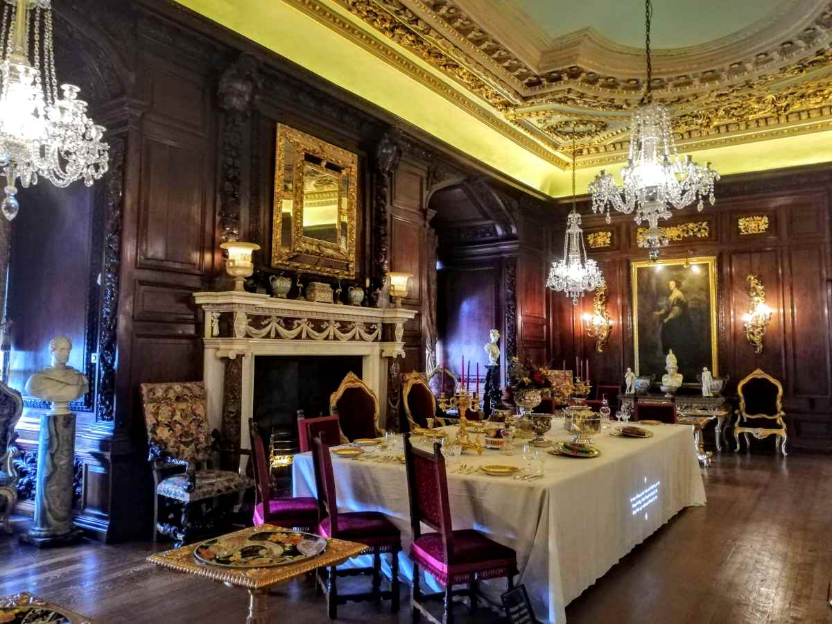 A formal dining room features wood-paneled walls, crystal chandeliers, and a long table set for a meal.