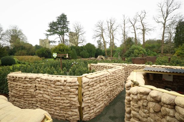 A sandbag wall structure sits in a garden area with a stone building visible in the background.
