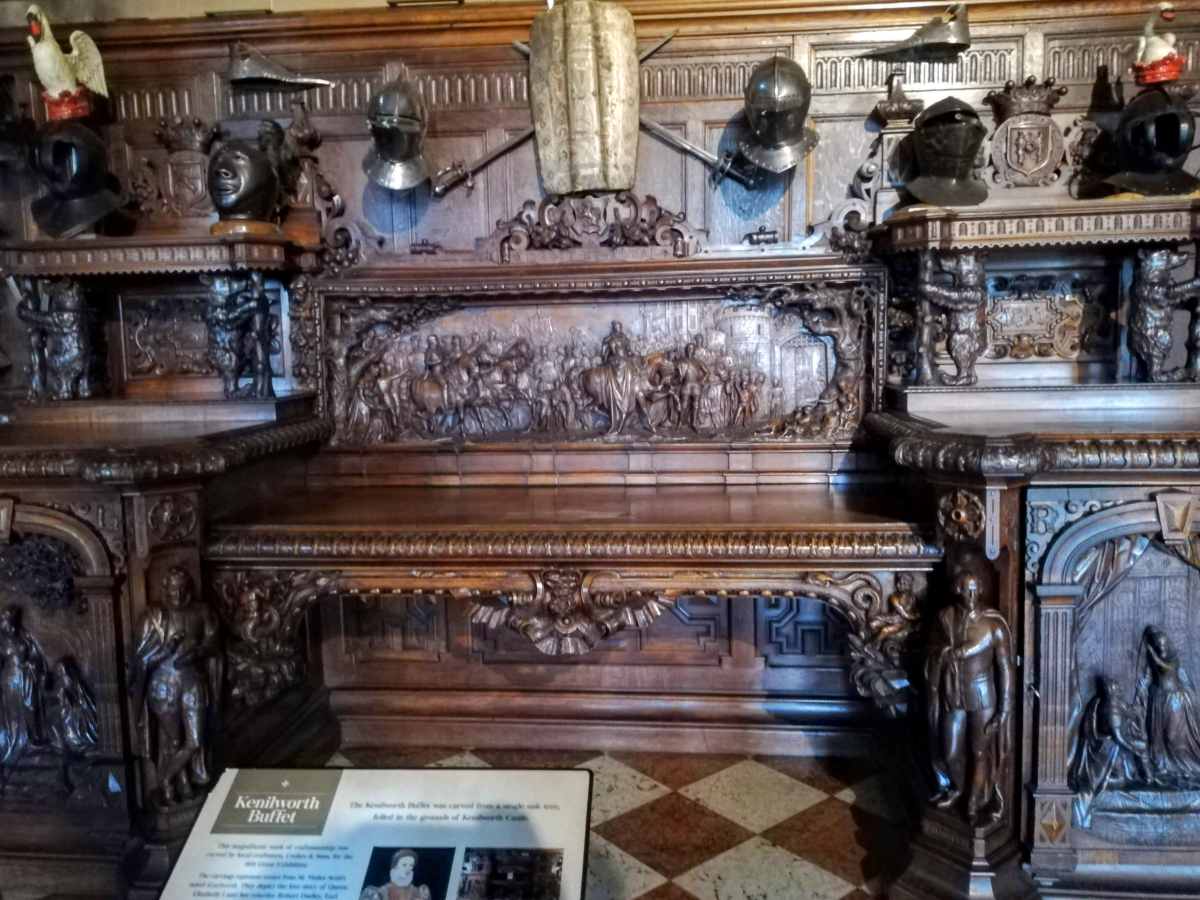 An ornate, carved wooden buffet is decorated with metal helmets and swords, with an informational sign in the foreground.