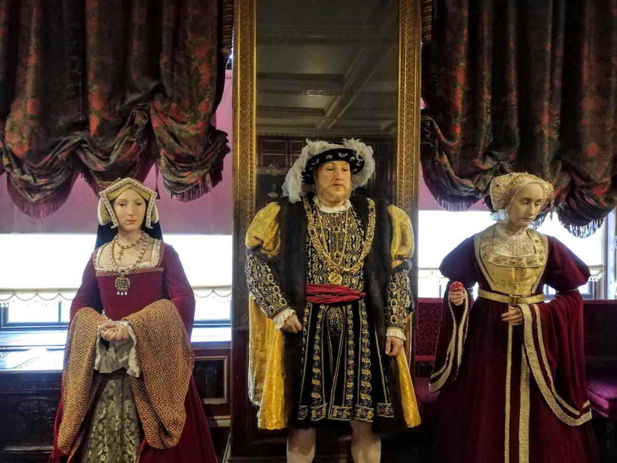 Three mannequins dressed in historical Tudor-style clothing stand in front of a large mirror and draped curtains.