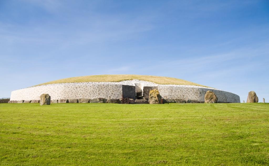 A grassy mound with a white stone wall and a central entrance sits in a field under a clear blue sky.