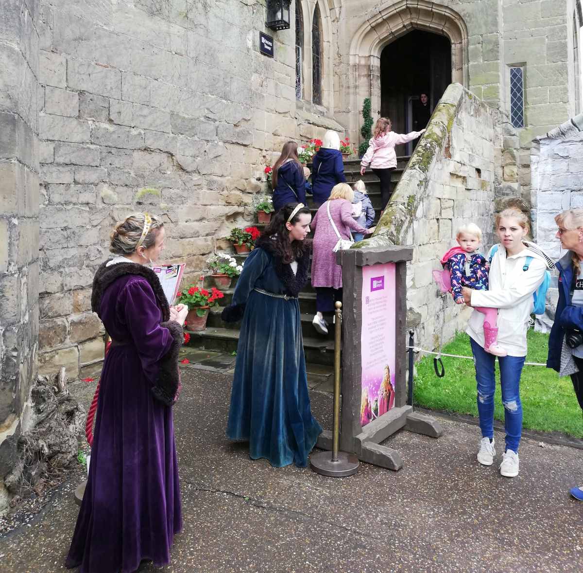 People in costumes and casual clothing stand near stone steps leading to a building entrance with a sign.