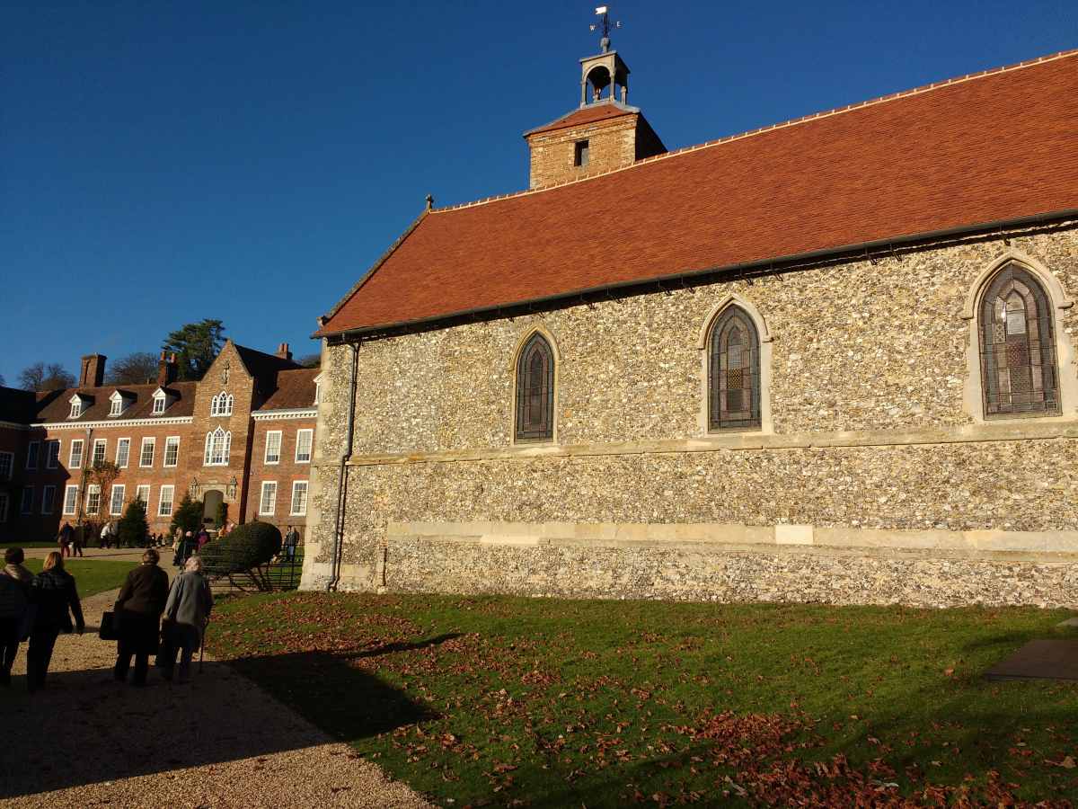 A stone church with a red tiled roof stands beside a brick building as people walk on a path in the foreground.