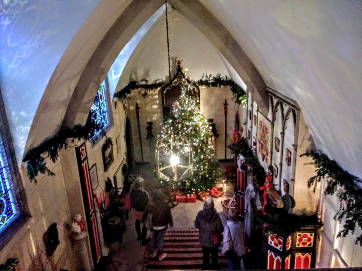 People walk down stairs toward a lit Christmas tree inside a stone building with vaulted ceilings and stained glass.