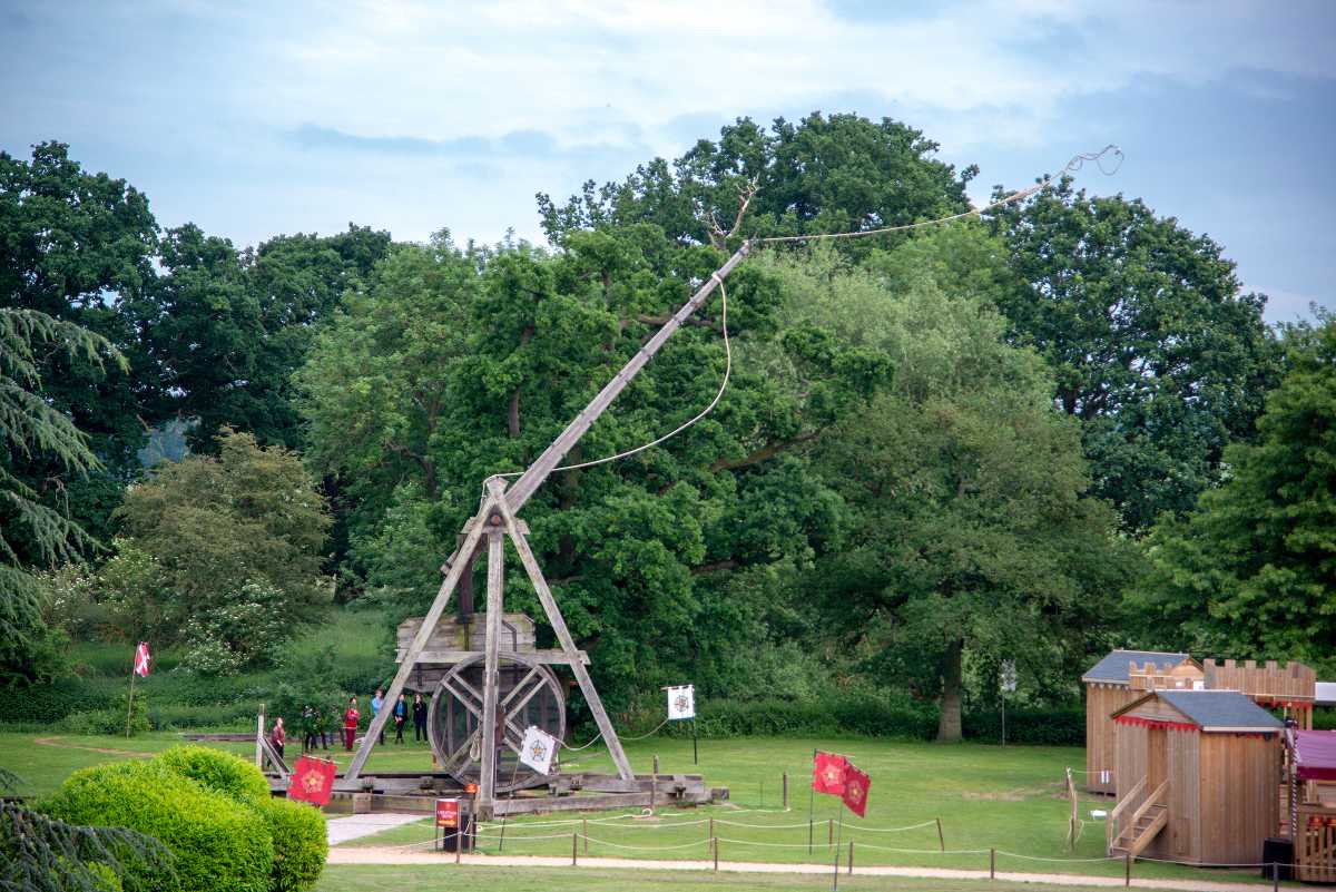 A large wooden trebuchet stands in a grassy field surrounded by trees, with small wooden structures and flags nearby.
