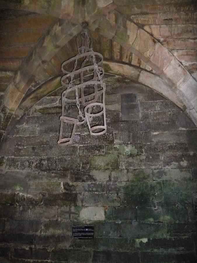 A metal cage hangs from a stone vaulted ceiling against a dark, weathered brick wall.