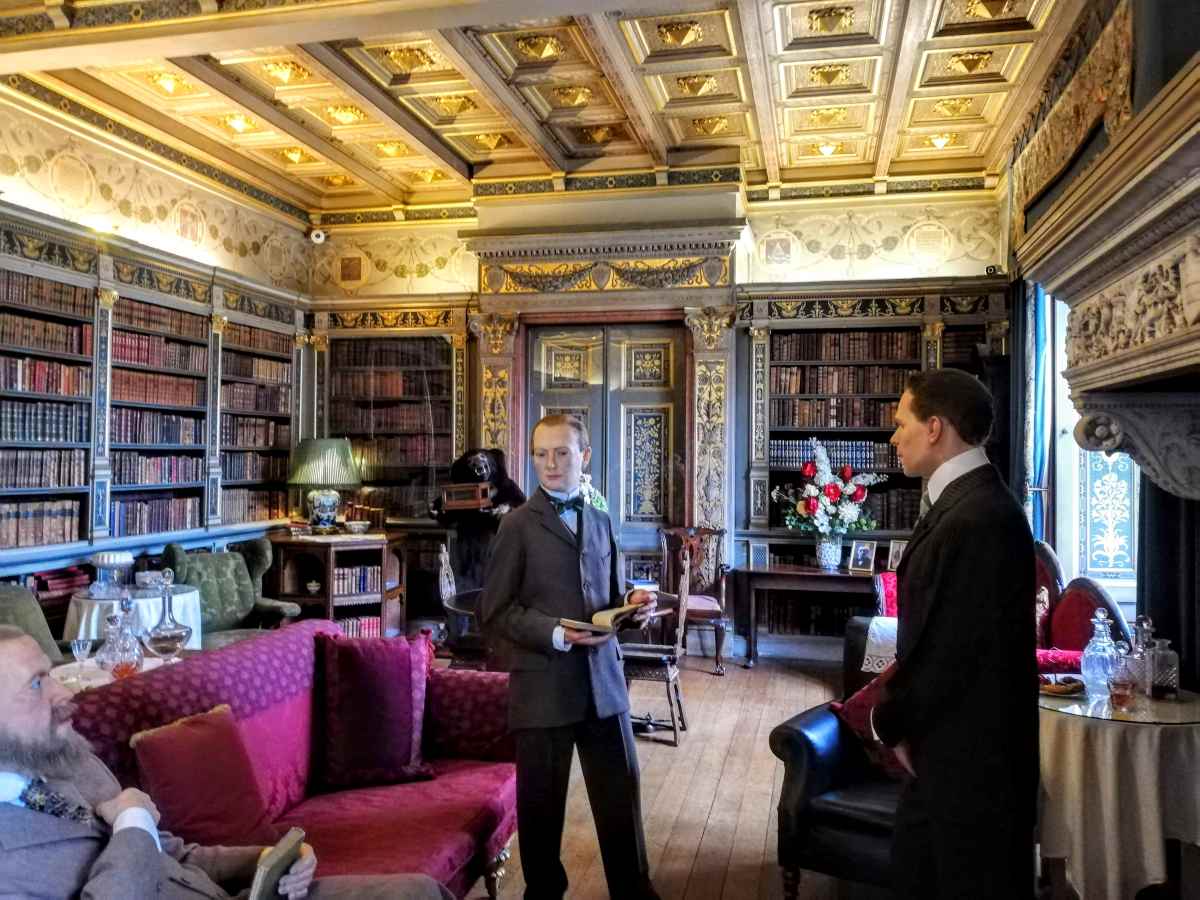 Three figures in period clothing stand in a library with floor-to-ceiling bookshelves and an ornate coffered ceiling.