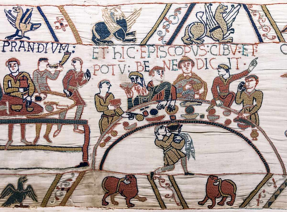 An embroidered tapestry depicts figures dining at tables with Latin text and decorative animal motifs above and below.