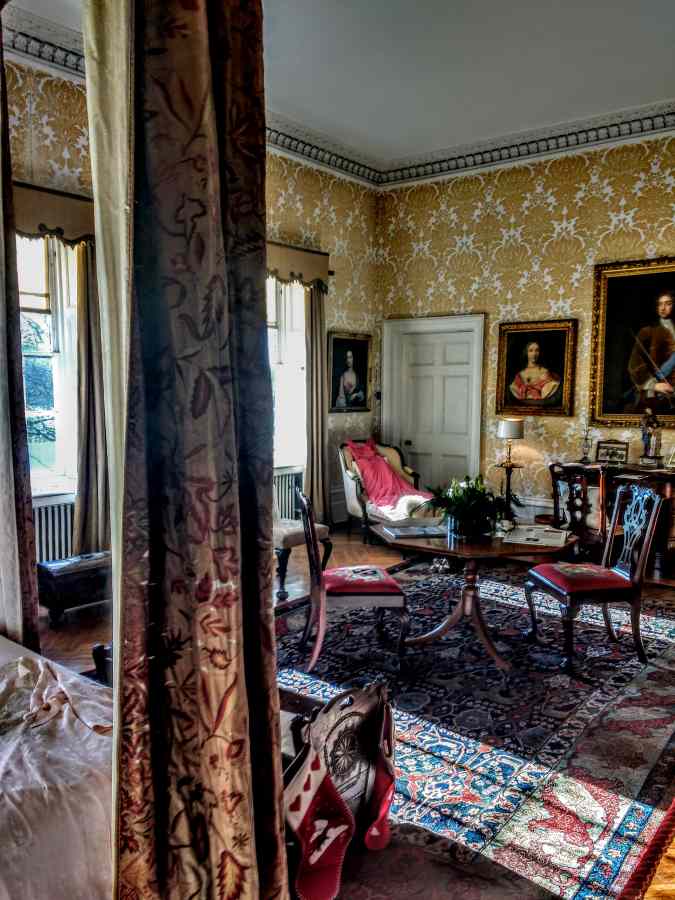 A room with patterned yellow wallpaper, framed portraits, a central table, and chairs on a patterned rug.