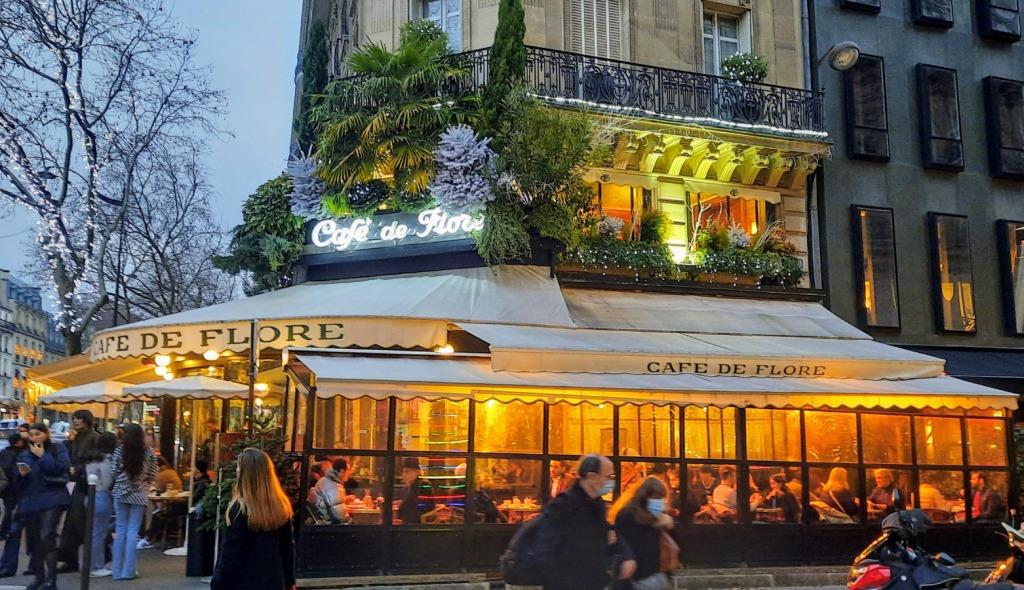 People walk past the Cafe de Flore, which features a lit, enclosed patio decorated with greenery and festive lights.