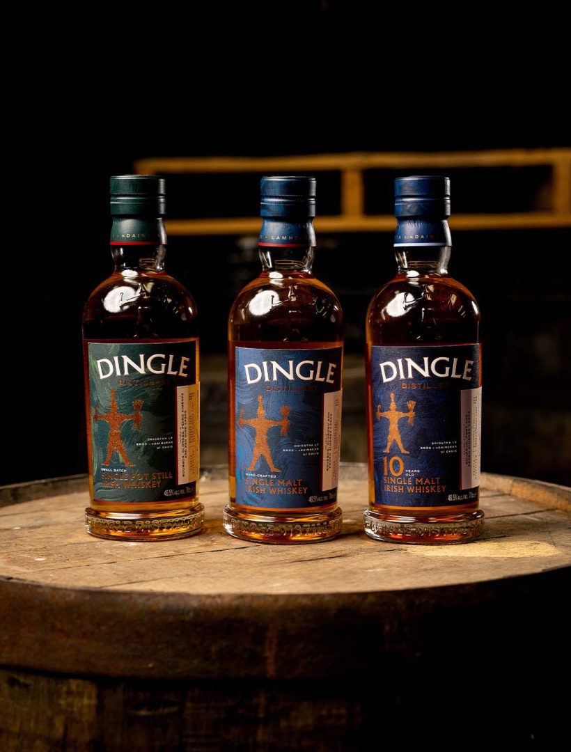Three bottles of Dingle Irish whiskey stand on a wooden barrel against a dark background.
