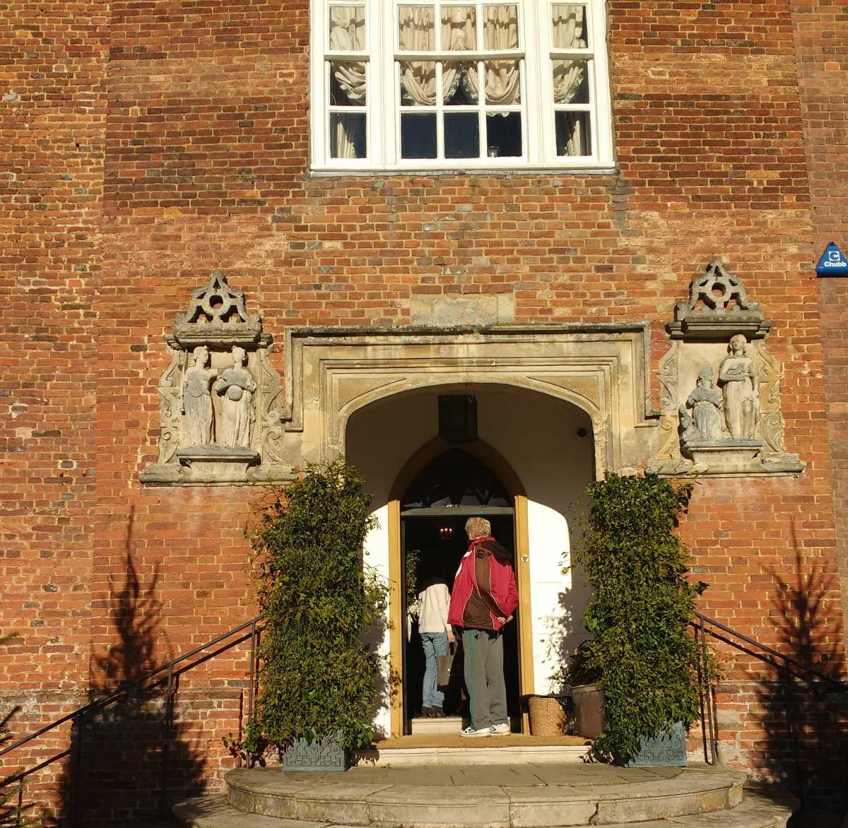 Two people enter a brick building through an arched doorway flanked by stone carvings and potted plants.