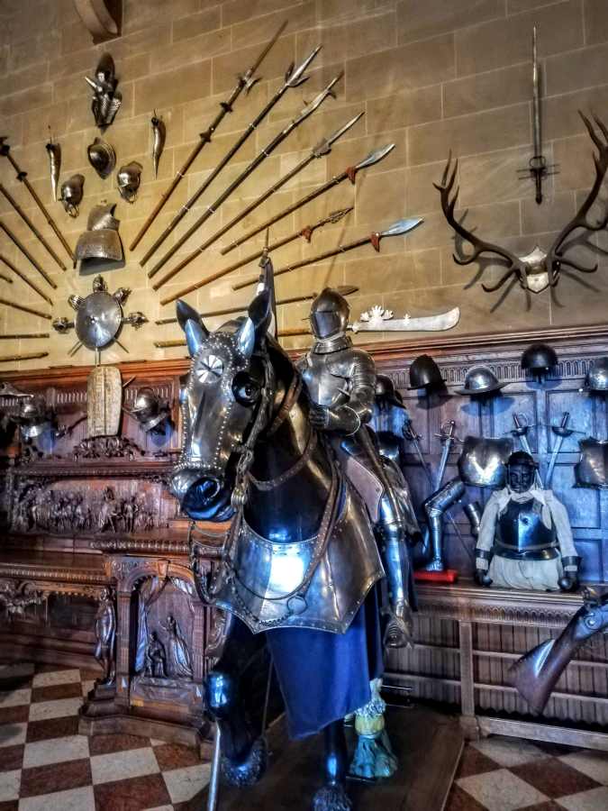 A suit of armor on a horse stands before a stone wall decorated with antique polearms, helmets, and a deer antler mount.